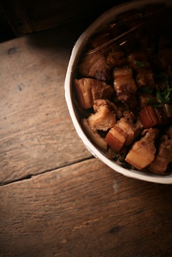 A bowl filled with chunks of pork belly, possibly braised, is placed on a rustic wooden table. The meat appears succulent and is garnished with green herbs, creating a homely and appetizing presentation.