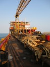 A large industrial platform on the sea with a crane structure and various equipment. Thick coils of rope are neatly arranged on wooden pallets, and yellow railings run along the edge. The platform has various levels of red and yellow-painted sections and appears to have operational buildings or control rooms at the rear. The sun casts long shadows, indicating it's either early morning or late afternoon.