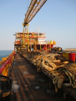 A large industrial platform on the sea with a crane structure and various equipment. Thick coils of rope are neatly arranged on wooden pallets, and yellow railings run along the edge. The platform has various levels of red and yellow-painted sections and appears to have operational buildings or control rooms at the rear. The sun casts long shadows, indicating it's either early morning or late afternoon.