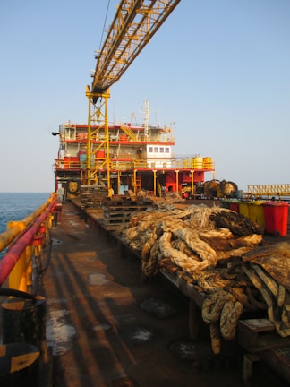 A large industrial platform on the sea with a crane structure and various equipment. Thick coils of rope are neatly arranged on wooden pallets, and yellow railings run along the edge. The platform has various levels of red and yellow-painted sections and appears to have operational buildings or control rooms at the rear. The sun casts long shadows, indicating it's either early morning or late afternoon.