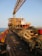 A large industrial platform on the sea with a crane structure and various equipment. Thick coils of rope are neatly arranged on wooden pallets, and yellow railings run along the edge. The platform has various levels of red and yellow-painted sections and appears to have operational buildings or control rooms at the rear. The sun casts long shadows, indicating it's either early morning or late afternoon.