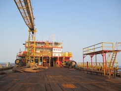 An offshore oil rig platform with large industrial structures and equipment visible. The metal framework is prominently yellow and red, with an expansive wooden deck in the foreground. The scene is set against a clear blue sky, suggesting a calm atmosphere.