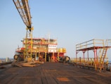 An offshore oil rig platform with large industrial structures and equipment visible. The metal framework is prominently yellow and red, with an expansive wooden deck in the foreground. The scene is set against a clear blue sky, suggesting a calm atmosphere.