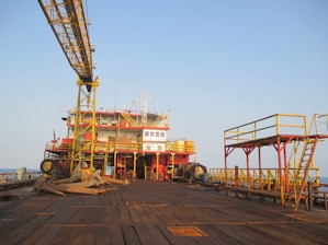 An offshore oil rig platform with large industrial structures and equipment visible. The metal framework is prominently yellow and red, with an expansive wooden deck in the foreground. The scene is set against a clear blue sky, suggesting a calm atmosphere.