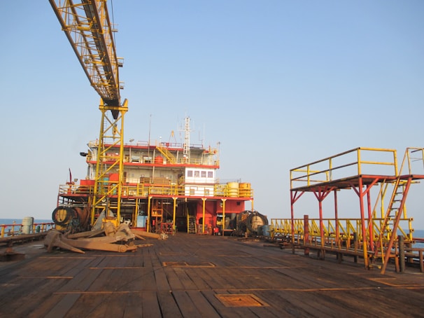 An offshore oil rig platform with large industrial structures and equipment visible. The metal framework is prominently yellow and red, with an expansive wooden deck in the foreground. The scene is set against a clear blue sky, suggesting a calm atmosphere.