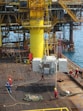 A group of workers in helmets and protective gear are maneuvering large industrial equipment on an offshore oil rig. The structure features prominent yellow piping and scaffolding, with the ocean visible in the background. The scene depicts a busy, industrial environment.