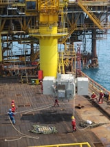 A group of workers in helmets and protective gear are maneuvering large industrial equipment on an offshore oil rig. The structure features prominent yellow piping and scaffolding, with the ocean visible in the background. The scene depicts a busy, industrial environment.