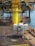 A group of workers in helmets and protective gear are maneuvering large industrial equipment on an offshore oil rig. The structure features prominent yellow piping and scaffolding, with the ocean visible in the background. The scene depicts a busy, industrial environment.