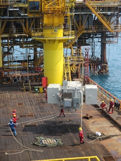 A group of workers in helmets and protective gear are maneuvering large industrial equipment on an offshore oil rig. The structure features prominent yellow piping and scaffolding, with the ocean visible in the background. The scene depicts a busy, industrial environment.