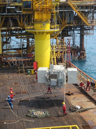 A group of workers in helmets and protective gear are maneuvering large industrial equipment on an offshore oil rig. The structure features prominent yellow piping and scaffolding, with the ocean visible in the background. The scene depicts a busy, industrial environment.