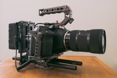 A professional camera setup with a large lens and a sturdy rig, placed on a wooden surface. The setup includes a handle grip on top and an external battery attached to the rear. The camera appears equipped for video work and has a robust and industrial design.