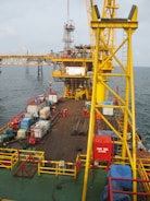 A large offshore platform with a yellow steel framework extends over the water. The site includes a deck filled with various containers, pipes, and equipment. Two workers in red outfits are visible on the platform deck. A drilling rig is centrally positioned, and there is a flame burning in the background, indicating the flaring of gas.