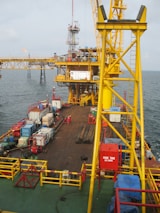 A large offshore platform with a yellow steel framework extends over the water. The site includes a deck filled with various containers, pipes, and equipment. Two workers in red outfits are visible on the platform deck. A drilling rig is centrally positioned, and there is a flame burning in the background, indicating the flaring of gas.