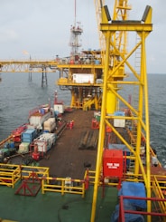 A large offshore platform with a yellow steel framework extends over the water. The site includes a deck filled with various containers, pipes, and equipment. Two workers in red outfits are visible on the platform deck. A drilling rig is centrally positioned, and there is a flame burning in the background, indicating the flaring of gas.