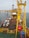 A large offshore platform with a yellow steel framework extends over the water. The site includes a deck filled with various containers, pipes, and equipment. Two workers in red outfits are visible on the platform deck. A drilling rig is centrally positioned, and there is a flame burning in the background, indicating the flaring of gas.