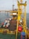A large offshore platform with a yellow steel framework extends over the water. The site includes a deck filled with various containers, pipes, and equipment. Two workers in red outfits are visible on the platform deck. A drilling rig is centrally positioned, and there is a flame burning in the background, indicating the flaring of gas.