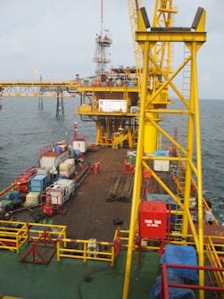 A large offshore platform with a yellow steel framework extends over the water. The site includes a deck filled with various containers, pipes, and equipment. Two workers in red outfits are visible on the platform deck. A drilling rig is centrally positioned, and there is a flame burning in the background, indicating the flaring of gas.