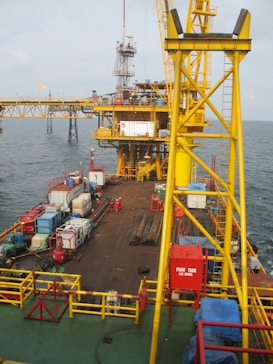 A large offshore platform with a yellow steel framework extends over the water. The site includes a deck filled with various containers, pipes, and equipment. Two workers in red outfits are visible on the platform deck. A drilling rig is centrally positioned, and there is a flame burning in the background, indicating the flaring of gas.