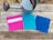 A rustic wooden surface features a metal watering can with a long spout and three colorful crochet washcloths. The washcloths are neatly lined up, showcasing different colors and patterns: vibrant pink with a white stripe, bright turquoise, and deep blue.
