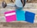 A rustic wooden surface features a metal watering can with a long spout and three colorful crochet washcloths. The washcloths are neatly lined up, showcasing different colors and patterns: vibrant pink with a white stripe, bright turquoise, and deep blue.