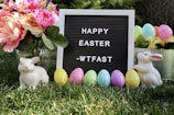 A spring scene featuring a letter board with the message 'Happy Easter' surrounded by colorful elements. On either side of the board, there are ceramic white rabbits, and a collection of pastel-colored Easter eggs placed on green grass. A bouquet of vibrant pink and orange flowers in a glass vase is positioned on the left, while a tin bucket filled with Easter eggs and flowers is on the right.