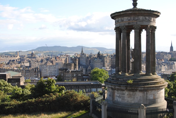 A neoclassical monument with tall columns stands in the foreground, overlooking a historical cityscape with a mix of old stone buildings and spires. Lush greenery surrounds the monument, and distant hills are visible under a partly cloudy sky.