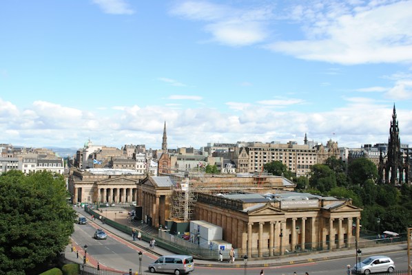 A cityscape featuring iconic neoclassical architecture, with several historic buildings forming a grand scene under a bright blue sky. A busy street with cars and pedestrians runs alongside a prominent gallery building, surrounded by lush greenery.