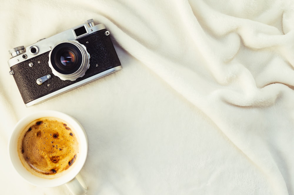 black and silver camera beside white ceramic mug on white textile