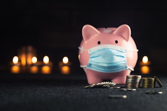 A pink piggy bank wearing a blue surgical mask is placed in the foreground, with several stacks and scattered coins nearby. In the background, there are blurred candlelights creating a warm, cozy ambiance.