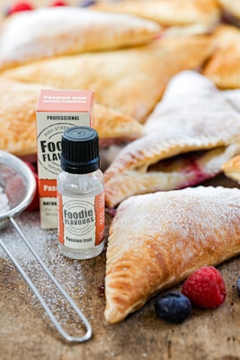 A close-up view of pastries sprinkled with powdered sugar, accompanied by small bottles of passion fruit flavoring. Fresh berries such as blueberries and raspberries are placed nearby, and a metal sieve is partially visible.