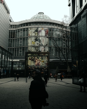 A modern urban setting with a glass building featuring large artistic murals. The building has a dome-like structure on top and wide glass windows. Several people are walking through the open space in front of the building, some in silhouette due to the lighting and time of day.