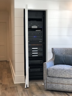 A neatly organized server rack is positioned within a recessed area of a room. The rack holds various electronic equipment, including amplifiers, switches, and other audio-visual components. The room features a wooden floor and white paneled walls. A gray upholstered armchair is placed nearby, adding a homey touch to the setting.