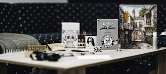 A wooden table with various items including a cup with the Starbucks logo, a painting of an architectural street scene, a small cartoonish feline painting, a book with Arabic text, a decorative box, and a few small figurines. The background features a polka-dotted sofa.