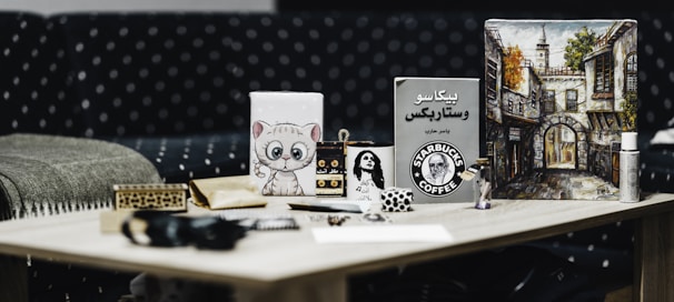 A wooden table with various items including a cup with the Starbucks logo, a painting of an architectural street scene, a small cartoonish feline painting, a book with Arabic text, a decorative box, and a few small figurines. The background features a polka-dotted sofa.