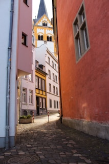 A narrow cobblestone alleyway flanked by buildings in pastel colors, leading towards a distant view of a Gothic-style church with a prominent spire and ornate facade. The sunlight casts shadows along the path, creating a peaceful and historic ambiance.