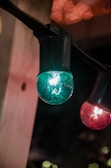 A close-up of decorative string lights suspended from a fixture. The image features a prominent turquoise globe-shaped bulb on the left and a red bulb on the right, with a soft warm glow in the background.