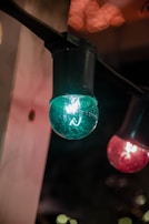 A close-up of decorative string lights suspended from a fixture. The image features a prominent turquoise globe-shaped bulb on the left and a red bulb on the right, with a soft warm glow in the background.
