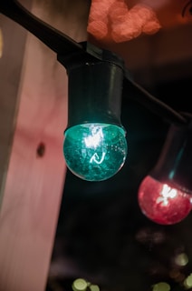 A close-up of decorative string lights suspended from a fixture. The image features a prominent turquoise globe-shaped bulb on the left and a red bulb on the right, with a soft warm glow in the background.