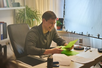A person is sitting at a desk in an office environment, smiling while looking at green sheets of paper. The workspace contains a plant, black office chair, stack of folders, glasses, and other office supplies. Soft natural lighting filters through a large window with sheer curtains.