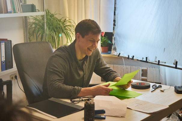 A person is sitting at a desk in an office environment, smiling while looking at green sheets of paper. The workspace contains a plant, black office chair, stack of folders, glasses, and other office supplies. Soft natural lighting filters through a large window with sheer curtains.