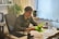 A person is sitting at a desk in an office environment, smiling while looking at green sheets of paper. The workspace contains a plant, black office chair, stack of folders, glasses, and other office supplies. Soft natural lighting filters through a large window with sheer curtains.
