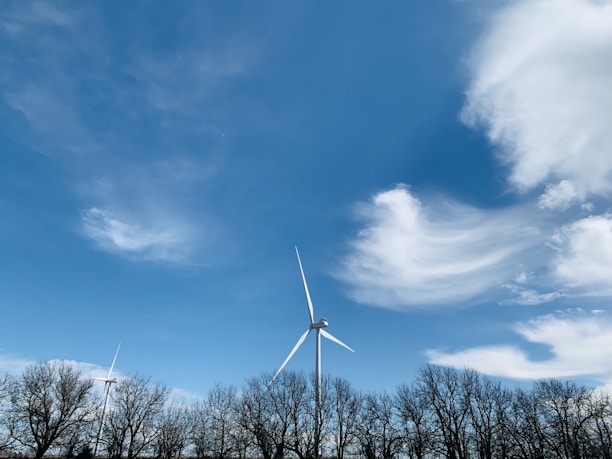 A wind turbine stands tall against a vivid blue sky with wispy white clouds. Bare trees line the horizon, indicating a winter or early spring setting.