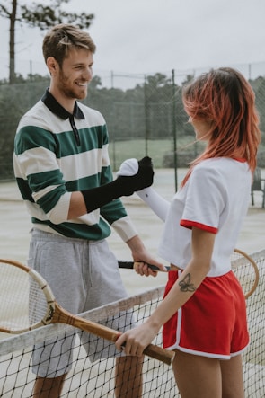 Two people are standing on a tennis court, each holding a tennis racket. They appear to be playfully interacting, perhaps engaged in a friendly competition or exchanging light-hearted banter.