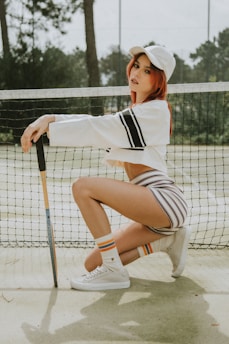 A person is squatting in front of a tennis net, wearing a white cap, a cropped long-sleeve white top with black stripes, striped shorts, and white sneakers with striped socks. The person is holding a tennis racket upright with one hand, and the background features a tennis court with a tall net and trees in the distance.