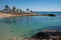 A scenic beach with calm turquoise waters bordered by a golden sandy shore and clusters of palm trees. Several people are enjoying the water and sunbathing on the beach. Rock formations are visible near the water's edge, adding texture to the serene landscape.