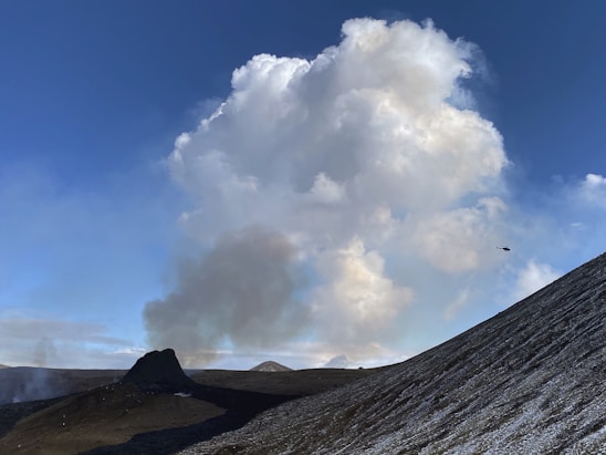 A large, fluffy cloud dominates the sky above a mountainous landscape. A plume of smoke rises from the ground, suggesting volcanic activity or a fire. A helicopter is visible in the distance, flying towards the smoke. The foreground features a steep, rocky slope.