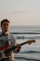 A musician plays an electric guitar in the foreground while in the background, a surfer rides a wave in the ocean. The focus is on the ocean and the surfer, with the setting sun casting a warm light across the scene.