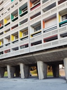 A section of a modernist residential building with a grid-like facade. The structure features unevenly sized rectangular windows with different colored panels, including green, yellow, red, and blue. The building is elevated on large concrete pillars, creating an open space underneath.