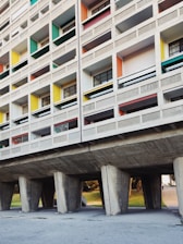 A section of a modernist residential building with a grid-like facade. The structure features unevenly sized rectangular windows with different colored panels, including green, yellow, red, and blue. The building is elevated on large concrete pillars, creating an open space underneath.