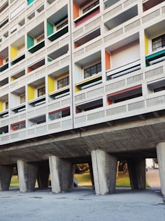 A section of a modernist residential building with a grid-like facade. The structure features unevenly sized rectangular windows with different colored panels, including green, yellow, red, and blue. The building is elevated on large concrete pillars, creating an open space underneath.